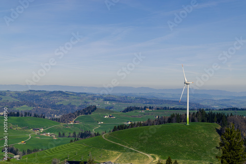 Wind turbine standing on a green hill above rolling countryside and scattered farms under a wide blue sky in a peaceful renewable energy landscape - entlebuch, lucerne, switzerland