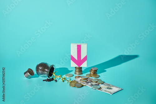 Fallen light bulb with sprout, euro coins and 100 dollar bill with down arrow sticker on turquoise background, failed mixed currency investment concept, copy space