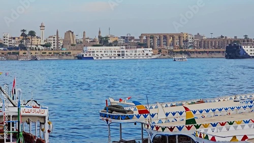 Colorful decorated local tourist ferry boats moored on Nile River at Luxor, Egypt, with cruise ships, Luxor Temple columns and city skyline visible across water.