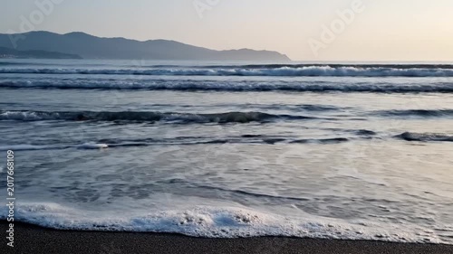 Serene coastal view with gentle ocean waves meeting a dark sandy beach. Distant mountains under a soft sky evoke natural tranquility and peaceful contemplation
