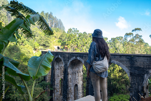 Woman traveler from behind looking at Nine Arch Bridge in Ella, Sri Lanka. Girl framed by banana leaves in front of historic railway viaduct.