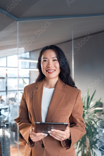 Vertical portrait middle-aged specialist business lady standing holding digital tablet at work in office, smiling at camera. Mature Asian professional manager woman leader entrepreneur using device