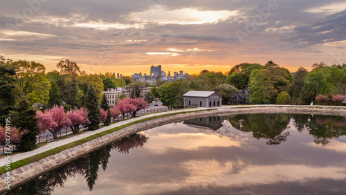 Aerial view of Brookline Reservoir with its stone gatehouse and blooming cherry blossoms against the distant Boston skyline at sunrise. Brookline, Massachusetts, United States.