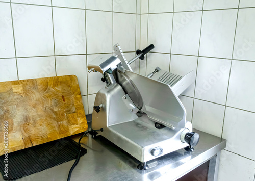 Electric deli meat slicer and wooden cutting board on a commercial kitchen counter, kitchen, equipment, blade, electric, metal, wooden, board, counter, prep table, tiles, white, professional, butcher,