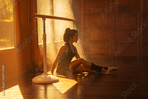 Ballerina in black sportswear practicing at a barre in a sunlit wooden studio.