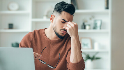Man sitting at a desk in a bright room shows signs of stress while holding his glasses. He is focused on work, with his hand on his forehead. Books and plants can be seen in the background.