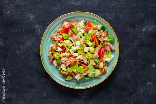 Cobb salad with chicken, avocado, tomatoes, eggs and bacon served in a light blue ceramic bowl on a black table, top view