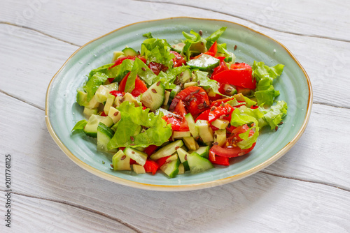 Fresh summer vegetable salad with cherry tomatoes, fresh cucumbers, avocado slices and lettuce, topped with flax seeds on a light blue ceramic plate on a white rustic wooden table close up.