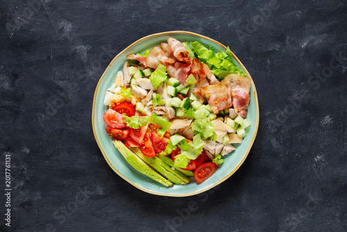 Cobb salad with chicken, avocado, tomatoes, eggs and bacon served in a light blue ceramic bowl on a black table, top view