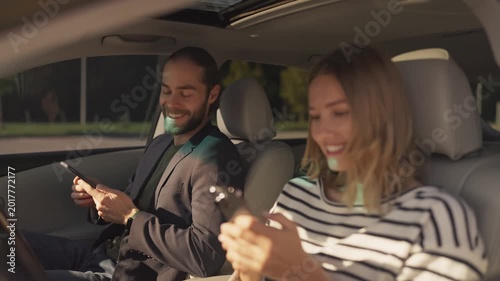 Smiling bearded man in blazer holding smartphone, sitting in driver’s seat and looking at woman in striped shirt beside him. Both appear cheerful, sharing moment inside parked car on sunny day.