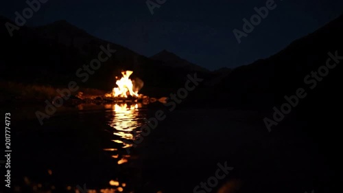 Campfire burning brightly on a calm lake at night, reflected in the dark water, surrounded by mountains