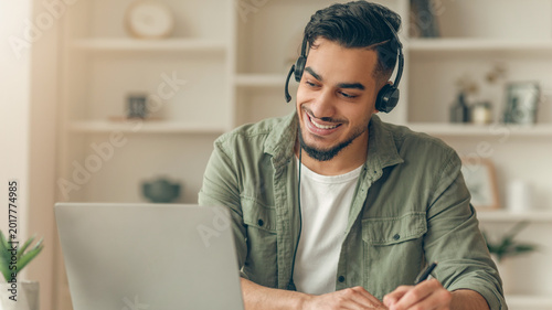 A young man sits at a desk in a bright room with a laptop and headset. He smiles while taking notes and participating in an online learning session. The scene shows focus and dedication.