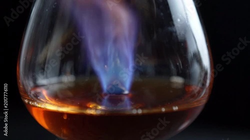 Flamb? technique, blue flames burning in a brandy snifter glass with amber liquid, dark background, close-up macro shot, culinary preparation, alcohol vaporization