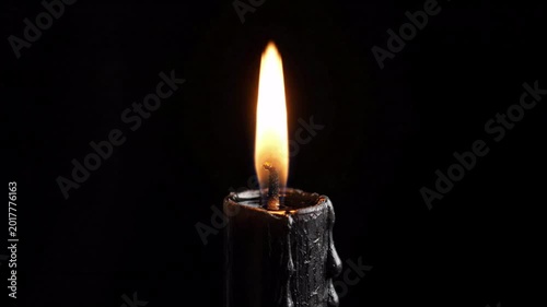 black wax candle burning brightly with a warm orange flame against a dark black background, dripping wax detail