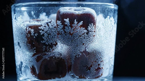 Close up of dark coffee ice cubes covered in frost and bubbles inside a clear glass with a dark background, refreshing drink element
