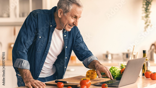 Φωτογραφία A man works in the kitchen, chopping vegetables while looking at a laptop