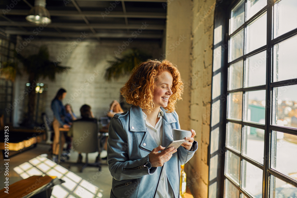 Fototapeta premium Smiling young woman with coffee and smartphone by office window in modern coworking space