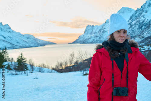 A traveler stands on snow in Norway, enjoying the view of mountains, lake, and sunset. The person wears a warm coat and prepares to capture the scene with a camera.