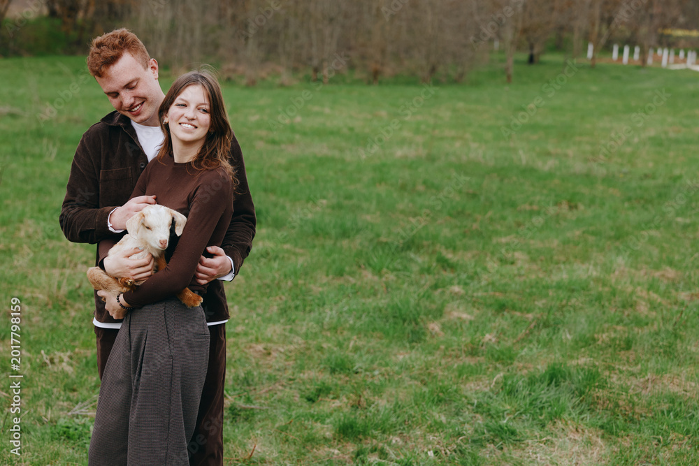 Naklejka premium Joyful couple outdoors with a goat kid, embracing in a grassy field, warm sun, relaxed pose, candid moment, natural colors, rural backdrop, simple happiness, wholesome lifestyle