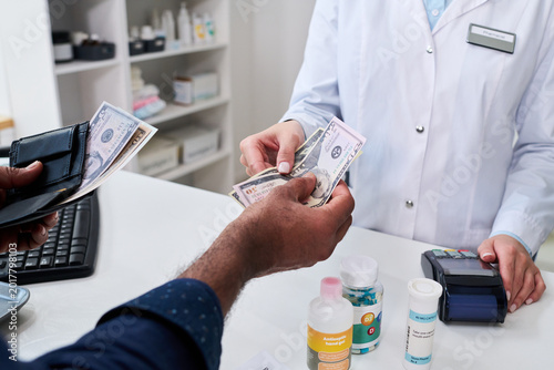 Close up of pharmacist receiving cash from customer at pharmacy counter, processing medication purchase and healthcare payment. Useful for insurance, prescription costs, retail care