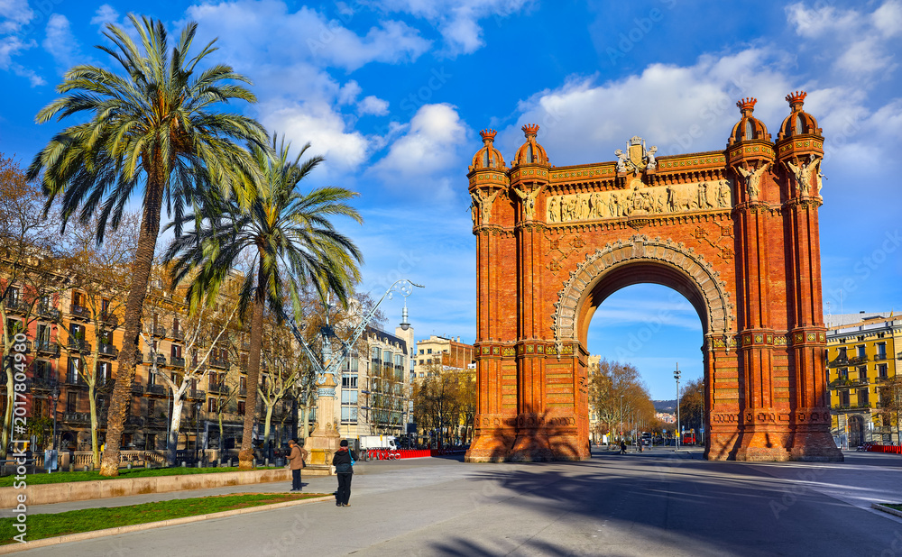 Fototapeta premium Barcelona, Catalonia, Spain. Triumphal Arch. Arc de Triomf at boulevard street. Alley with tropical palm trees. Early morning landscape with shadows and blue sky with clouds. Famous landmark.