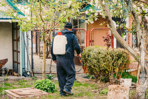 Man sprays plant in garden surrounded by blooming trees and house on a clear day