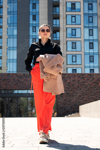Young fashionable woman with coat over arm posing in front of contemporary glass and brick building. Urban lifestyle and street fashion empowerment concept.
