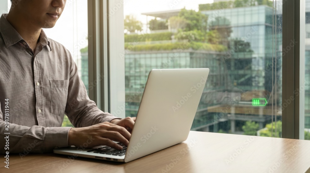 Fototapeta premium Man typing on a laptop near a window showing a green battery charging icon