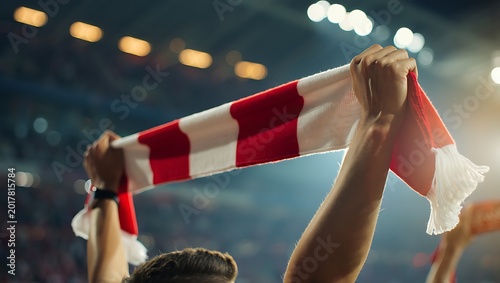 Excited soccer fans holding up a red and white scarf in a crowded stadium at night