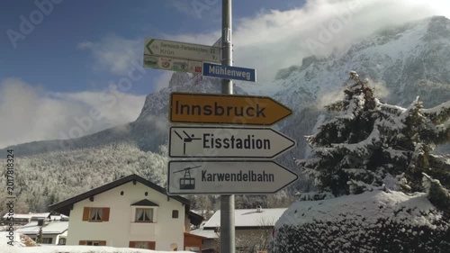 Road signs in Mittenwald, Bavaria, Germany, pointing toward Innsbruck and local destinations. Snow covered houses and navigation signs stand against Karwendelgebirge, with Westliche Karwendelspitze