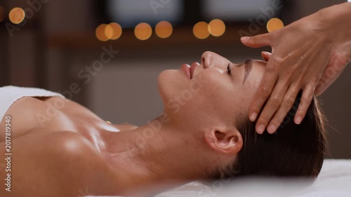 A woman with light skin receives a head massage in a spa setting. The atmosphere is warm with soft lighting. The focus is on the woman's peaceful expression and the therapist's gentle touch.