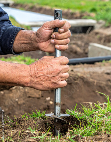 A field researcher's hands use a metal soil probe to extract a core sample from a grassy bank near an industrial water outlet.
