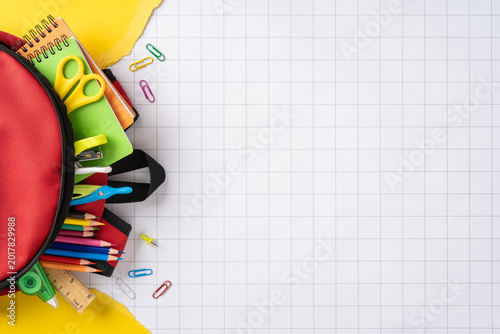 Back to school concept with red backpack full of colorful pencils, notebooks and supplies on graph paper