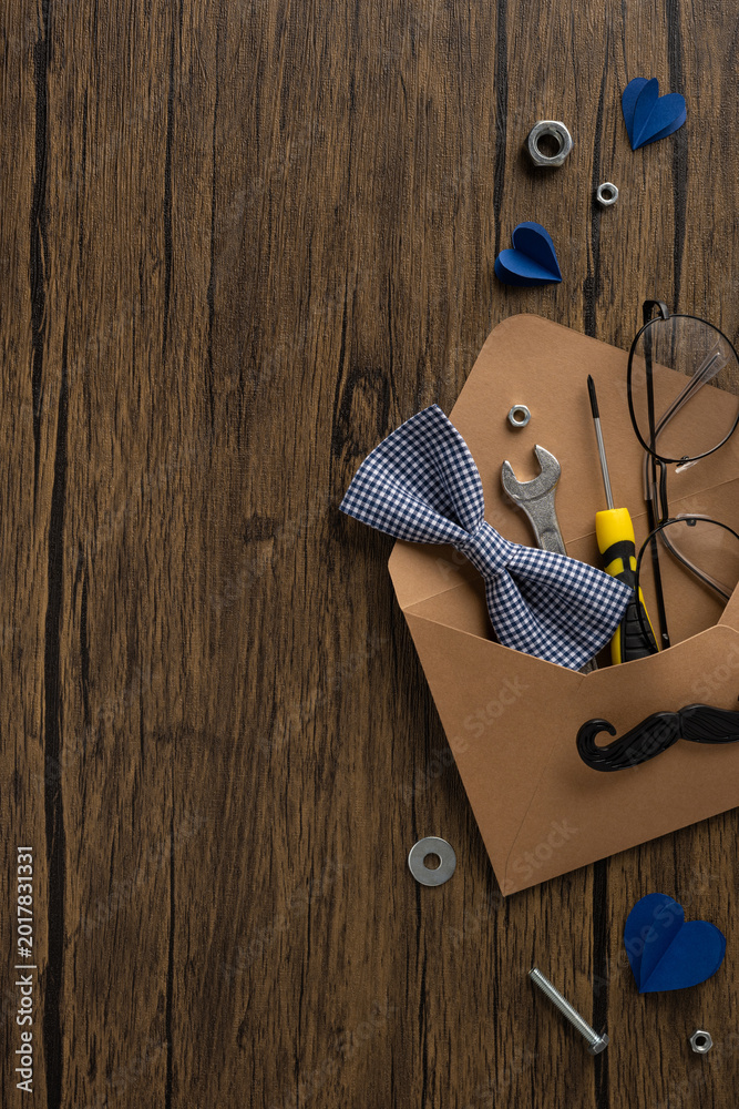 Fototapeta premium Happy Father's Day gift vertical envelope with tools, bow tie, glasses and paper hearts on wooden table