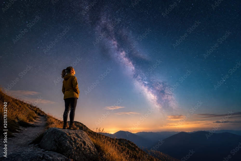 Fototapeta premium Person standing on mountain cliff under the Milky Way at sunrise with dramatic twilight sky and distant peaks