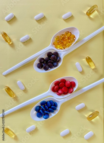 Creative flat lay composition with colorful capsules, vitamins, and fish oil supplements arranged in white ceramic spoons on a yellow background. Concept of healthy nutrition, dietary supplements