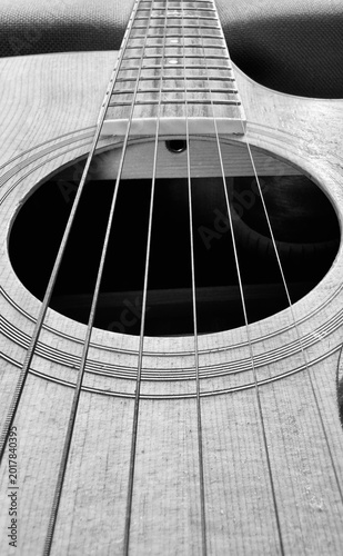Detailed black and white close-up photograph of acoustic guitar strings and sound hole viewed from the neck perspective. Artistic music background with selective focus highlighting instrument texture
