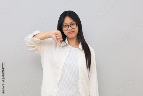 Cuadro en lienzo A young Asian woman with a displeased expression, wearing casual clothes, showing a thumbs down, a gesture of dislike, rejection, standing isolated on a white background