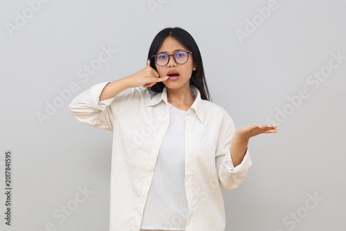 Photos A young Asian woman wearing casual clothes, making a phone gesture with her finger, looking confused and hesitant, wondering, standing alone on a white background