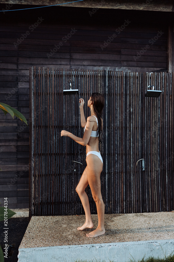 Obraz premium A woman in a white swimsuit walks beside an outdoor shower in a sunny setting, embracing a casual lifestyle moment. The scene captures everyday relaxation, personal care, and outdoor leisure,