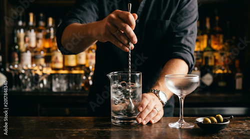 Hands of bartender stirring clear cocktail in mixing glass with long bar spoon beside coupe glass and small dish of olives on dark bar