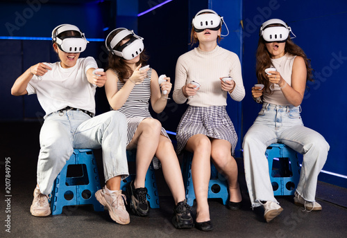 Fotografía Four young women sitting on stools and enjoying virtual reality gaming with VR headsets and controllers in modern gaming center, fully engaged in immersive experience