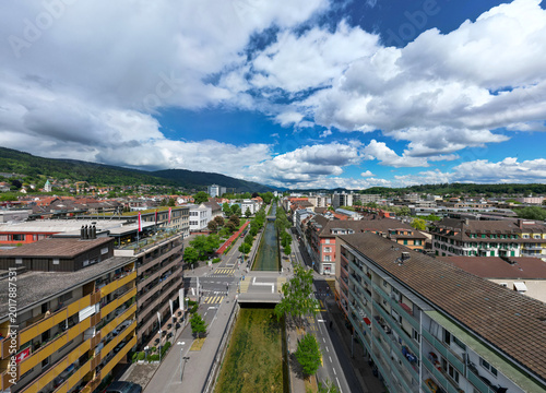 Aerial view of the Schuss canal flowing through the urban landscape with residential buildings and green hills under a cloudy sky in Biel/Bienne, Canton of Bern, Switzerland.