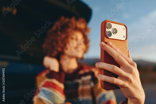 Woman lifestyle smartphone selfie outdoors, curly hair, fashion striped top, hand holding a phone case for a candid shot, blurred city background, daylight and warm tones, soft bokeh today