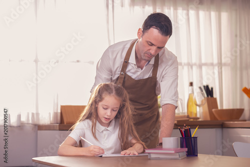 Daughter Sitting At Table Doing Homework As Father Cooks Meal. Father teach little girl how to draw the artwork while cooking dinner in a kitchen.
