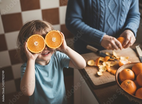 Child holding orange halves over eyes, person peeling oranges