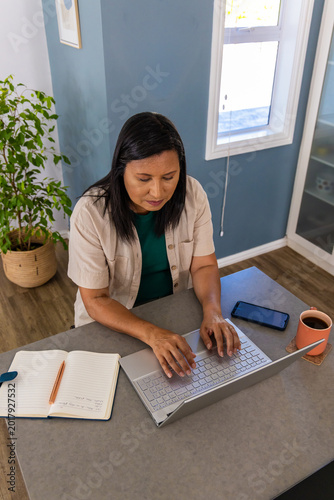 Mature woman sitting at home workspace typing on silver laptop with notebook smartphone and mug
