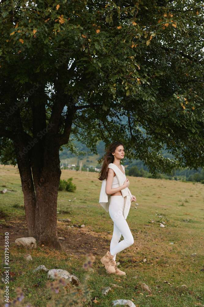 Obraz premium Woman lifestyle outdoors, standing near a tree in a sunlit meadow, wearing white outfit, relaxed pose, gentle breeze, natural colors, peaceful countryside.