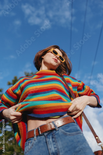 Foto Woman, young woman in a colorful striped sweater and yellow sunglasses, pulling up sweater to show midriff in jeans outdoors under bright blue sky, playful confident travel style