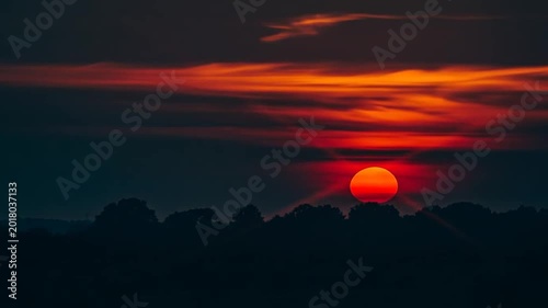 A vibrant orange sun sets behind a dark silhouette of trees at dusk